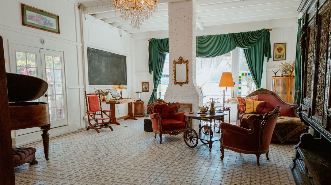 The Crescent House parlor set for gatherings, with a crystal chandelier, a white brick fireplace, green drapes, and Victorian furniture on the original tile