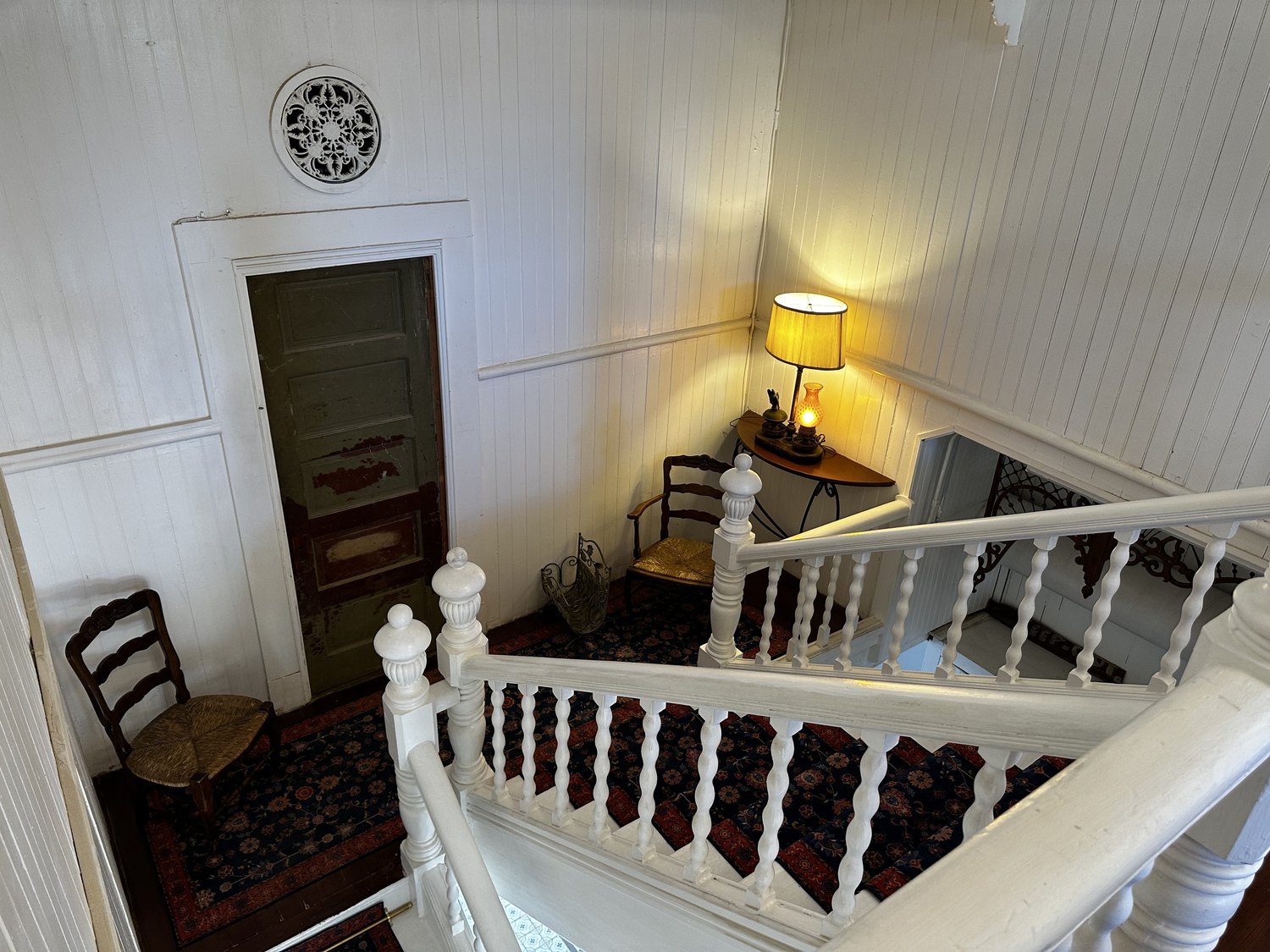 The landing between the two flights of stairs at The Crescent House, seen from above, with its white balustrade, patterned runner, and a lamp on a half-round table